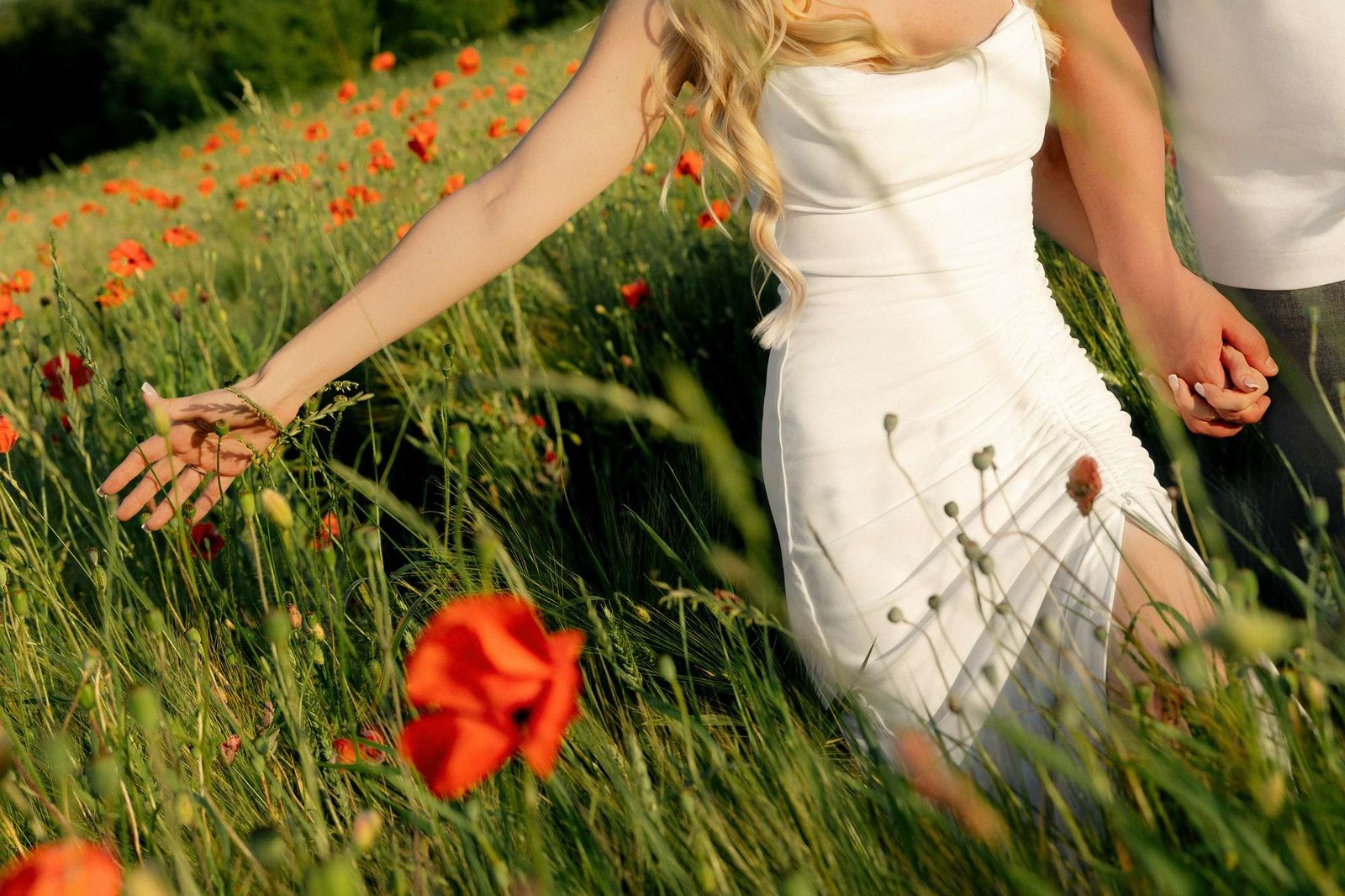 Bride-to-be walking through wildflowers, her hand brushing the grass, holding her partner's hand