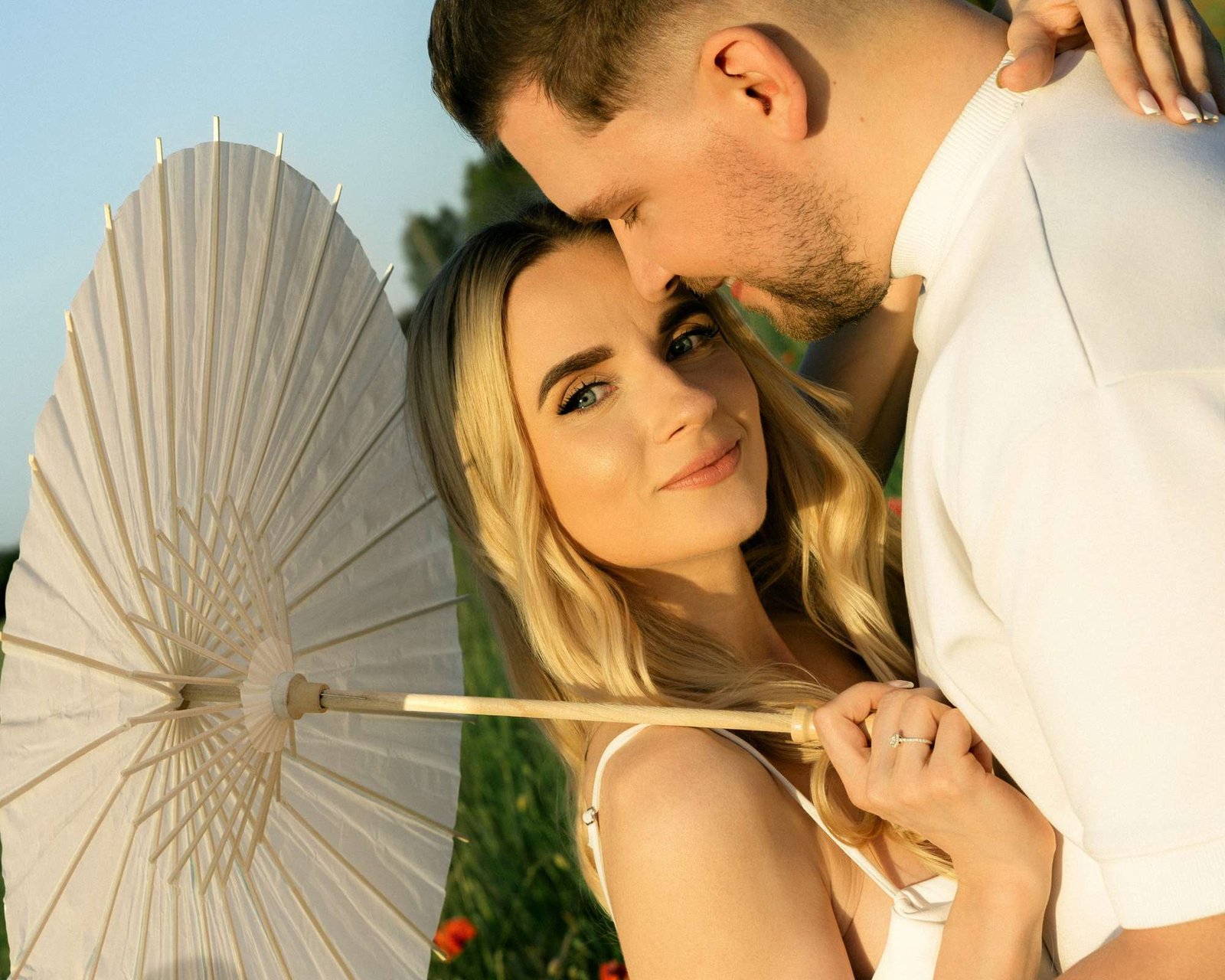 Man kissing his partner's forehead while she holds a white parasol in the meadow