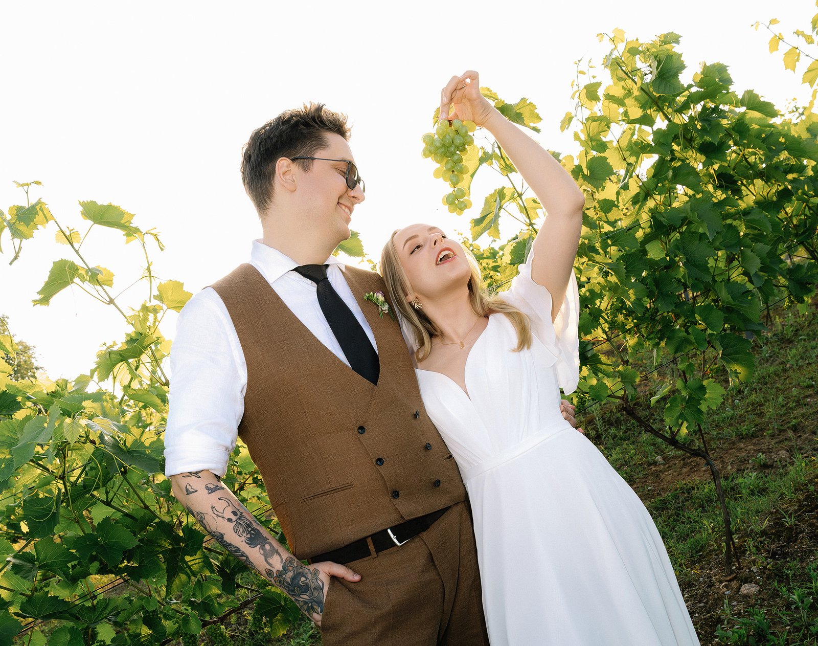 Bride laughing and trying to eat a bunch of grapes while the groom watches at a vineyard wedding in Italy